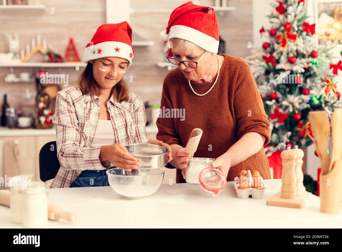 Mehrere Generationen Familie macht zusammen Cookies am weihnachtstag. Happy fröhlich freudig Teenager-Mädchen hilft älteren Frau Vorbereitung süßen buiscuits Winterferien tragen weihnachtsmütze zu feiern. Stockfoto