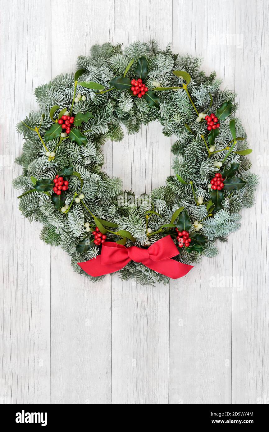 Weihnachten Stechpalme, Mistel & Schnee bedeckt Fichte Fi Winterkranz mit roter Schleife auf rustikalem Holz vor Tür Hintergrund. Traditionelles Thema für die Solstic Stockfoto