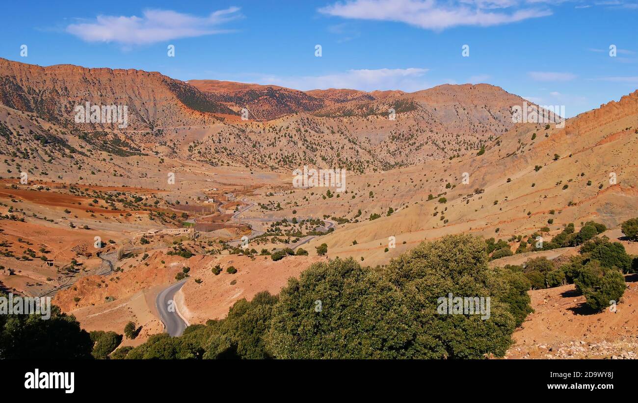 Schöner Panoramablick über ein spärliches Tal mit einer Straße, die zum Bergdorf Imilchil in den Altas Bergen, Marokko mit verstreuten Bäumen führt. Stockfoto