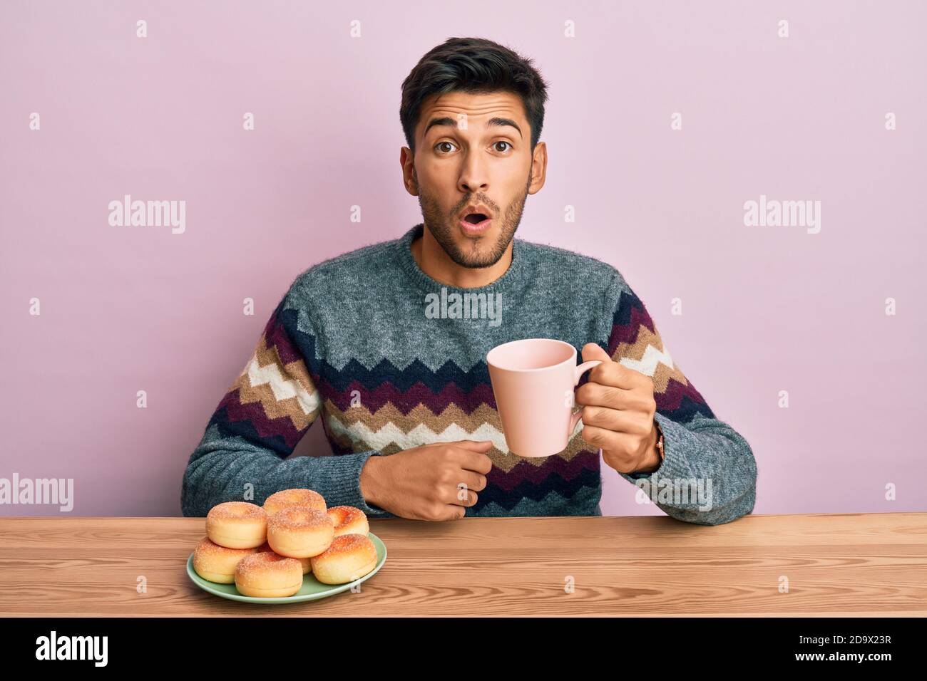 Junger gutaussehender Mann essen Donuts und trinken Kaffee verängstigt und erstaunt mit offenem Mund für Überraschung, Unglauben Gesicht Stockfoto