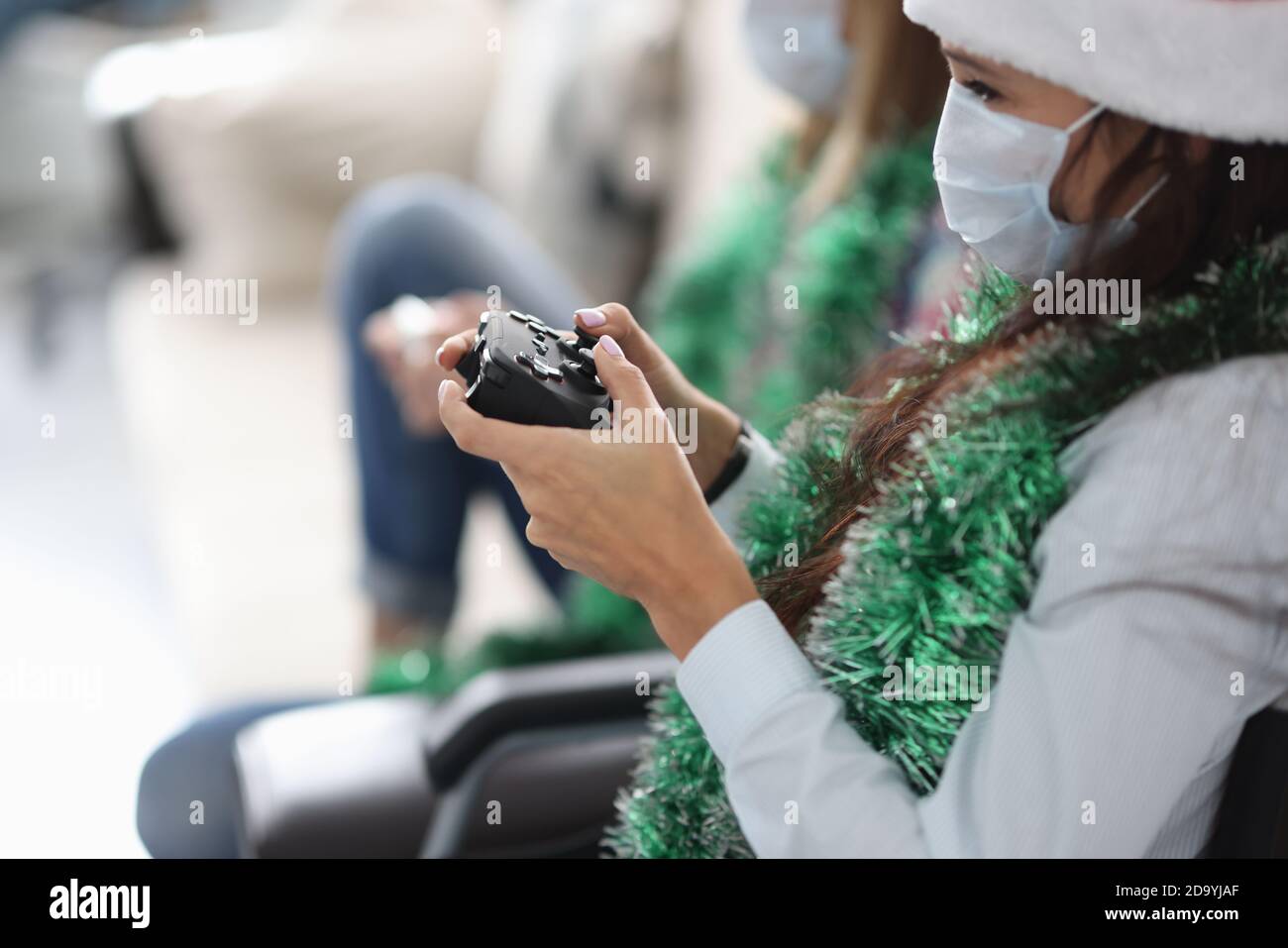 Zwei Freundinnen sitzen auf der Couch mit Spiel-Joysticks In santa claus Hüte und schützende medizinische Masken Stockfoto