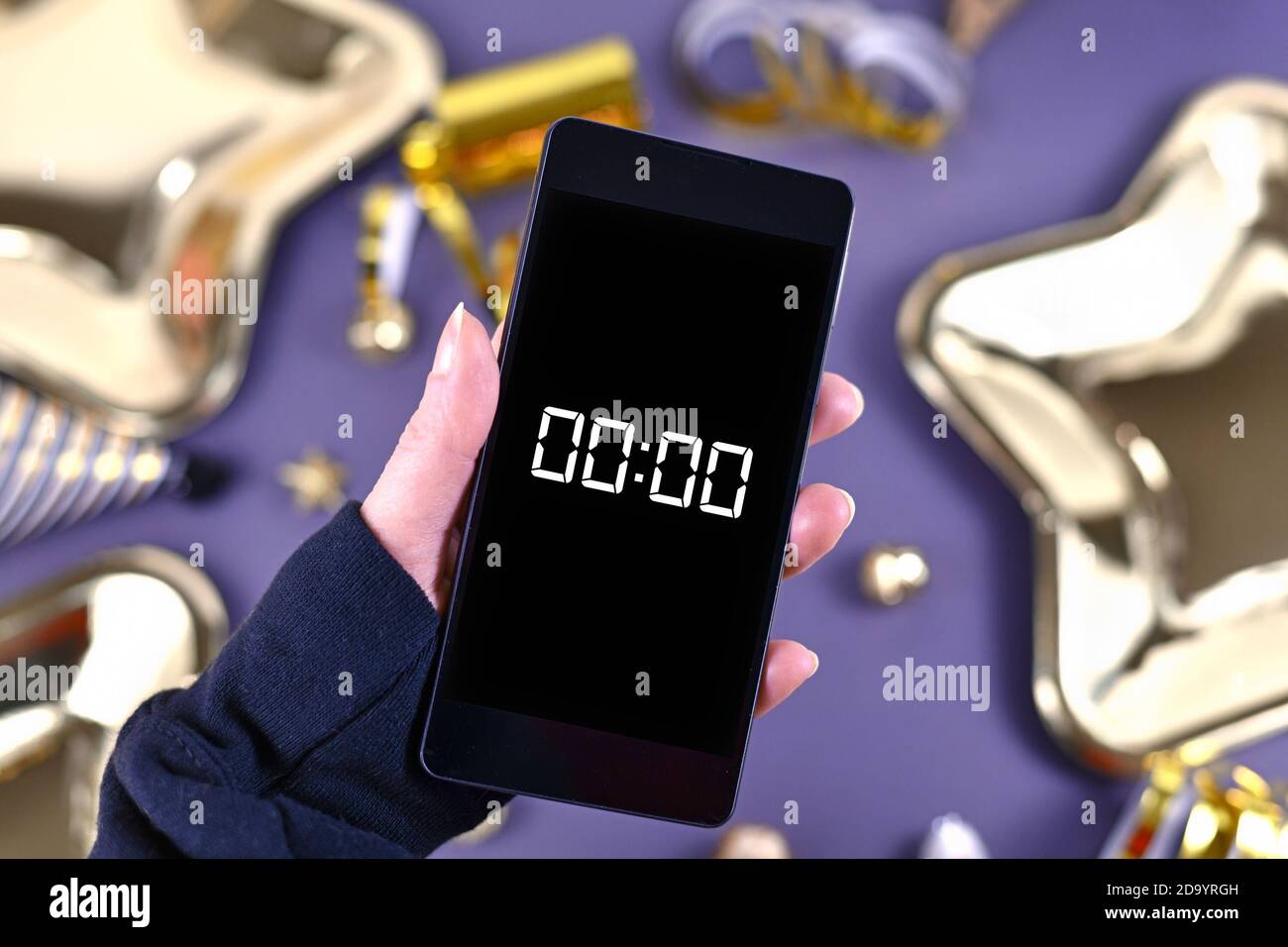 Silvester-Festkonzept mit Handgriff auf Smartphone Mit Timer Countdown bis Mitternacht vor Party-Elemente Im Hintergrund Stockfoto