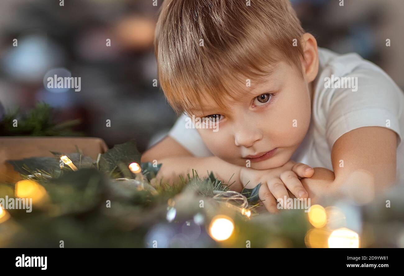 Ein schöner Junge von 4-5 Jahren sitzt oder steht in der Nähe des Weihnachtsbaums. Er träumt von einem Urlaub und Geschenke, macht einen Wunsch. Das Konzept des wi Stockfoto
