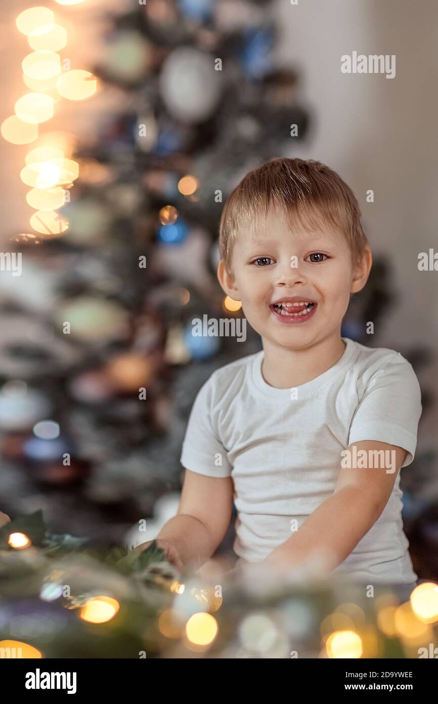 Ein schöner Junge von 4-5 Jahren sitzt oder steht in der Nähe des Weihnachtsbaums. Er träumt von einem Urlaub und Geschenke, macht einen Wunsch. Das Konzept des wi Stockfoto