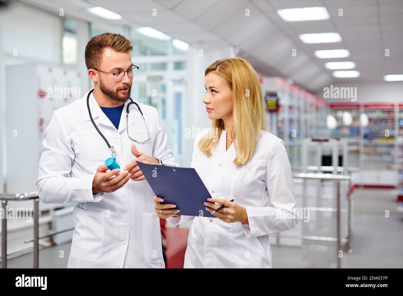 Zwei junge kaukasische Apotheker diskutieren Medikamente, mit einer digitalen Tablette und Dokumente. Mann und Frau in weißem medizinischen Kleid arbeiten in der Apotheke Stockfoto