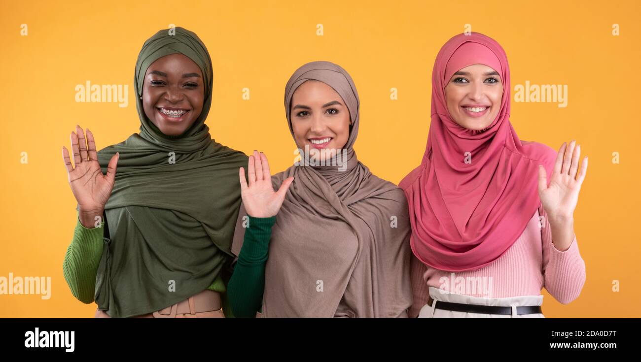 Drei Muslimische Damen Winken Hallo Geste Über Gelbem Hintergrund, Panorama Stockfoto
