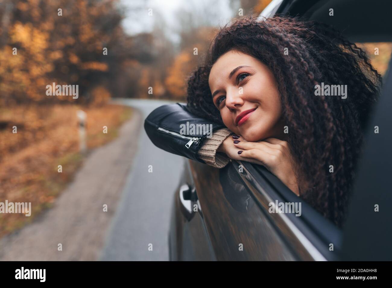 Junge afro Haar Frau reisen mit dem Auto auf wilden Wald Herbst Straße. Weiblicher Blick in geöffnetem Fenster von hinten sitzen mit glücklichem Lächeln Stockfoto
