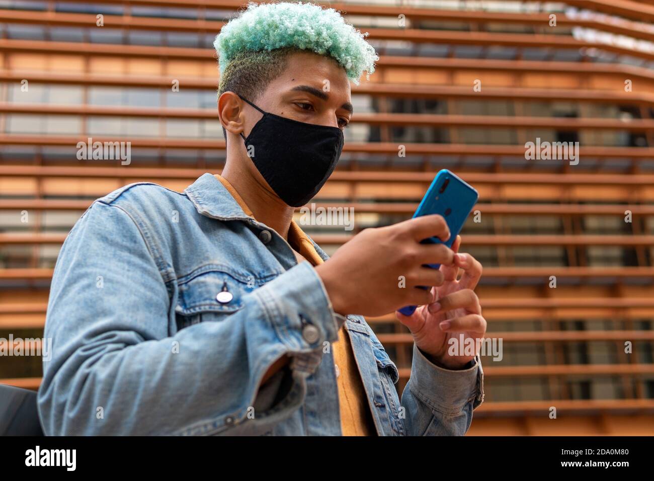 Niedriger Winkel der schweren ethnischen Männchen mit blauen Haaren und In Schutzmaske stehen im städtischen Bereich und Surfen im Internet Auf dem Smartphone während COVID 19 epide Stockfoto