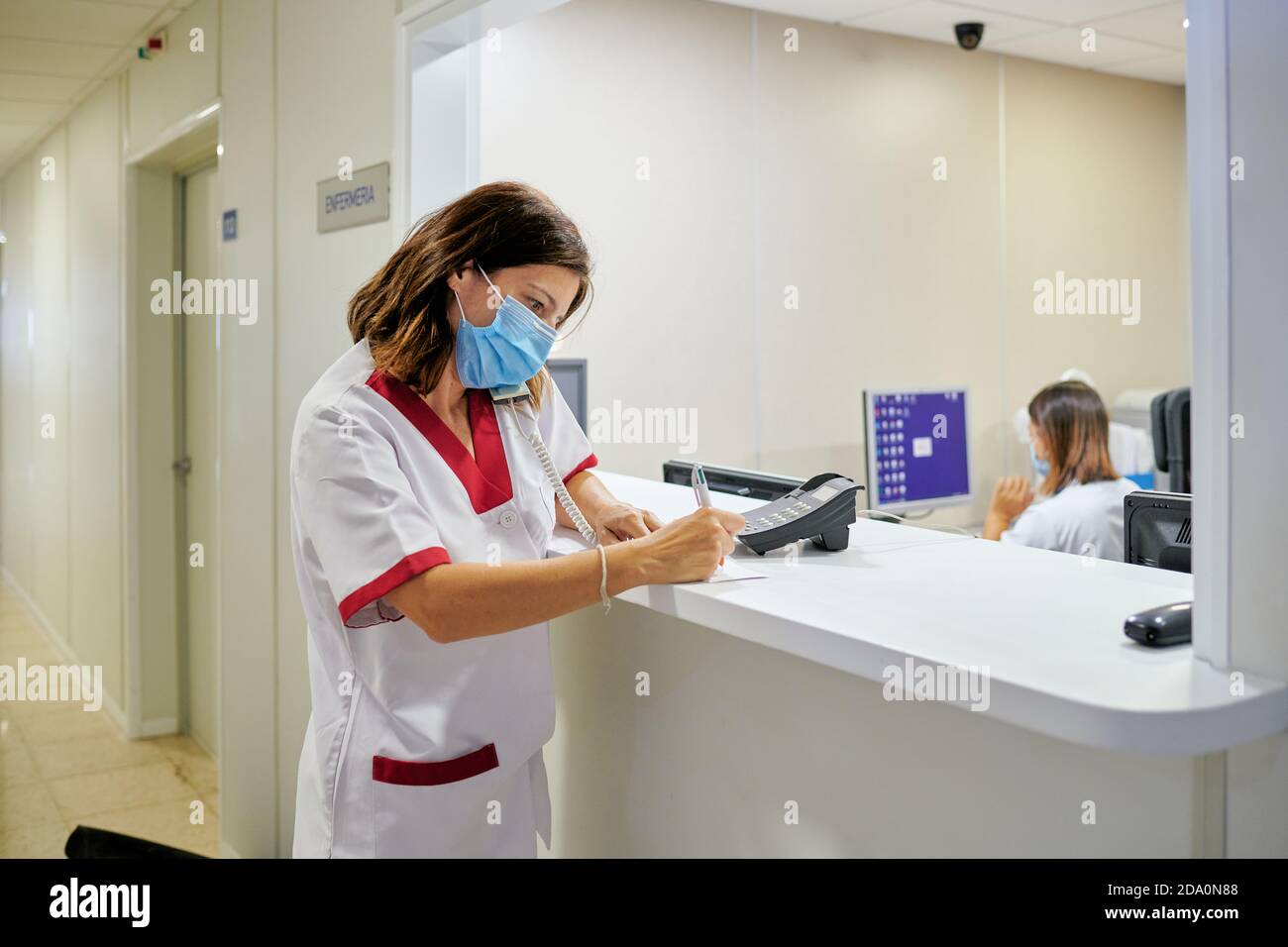 Konzentrierte weibliche Krankenschwester trägt weißen medizinischen Anzug und Gesichtsmaske Notizen in der klinischen Dokumentation machen, während man sich in der Nähe von modern befindet Krankenhausempfang Stockfoto