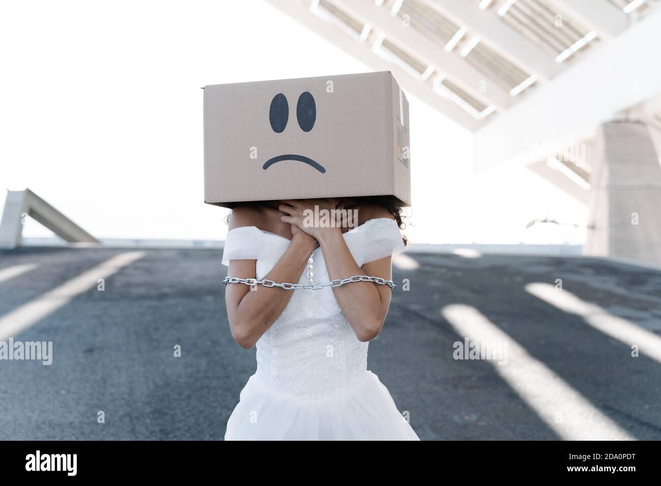 Anonyme Frau in weißem Brautkleid mit Metallkette Arme tragen Karton mit unglücklichen Smiley Stockfoto