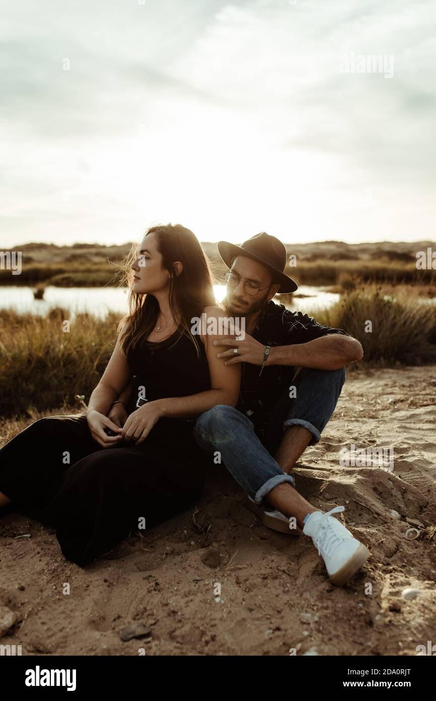 Junge liebevolle Paar in lässiger Kleidung sitzen zusammen Blick weg Auf der Küste des Stausees umgeben vom trockenen Gras in der Wiese Bei Sonnenlicht Stockfoto