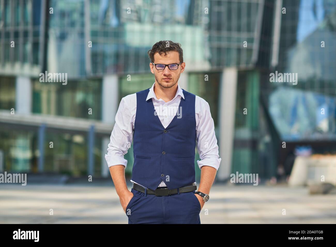 Junge ernsthafte Geschäftsmann in formelle Kleidung und Armbanduhr stehen mit Hände in den Taschen beim Betrachen der Kamera Stockfoto