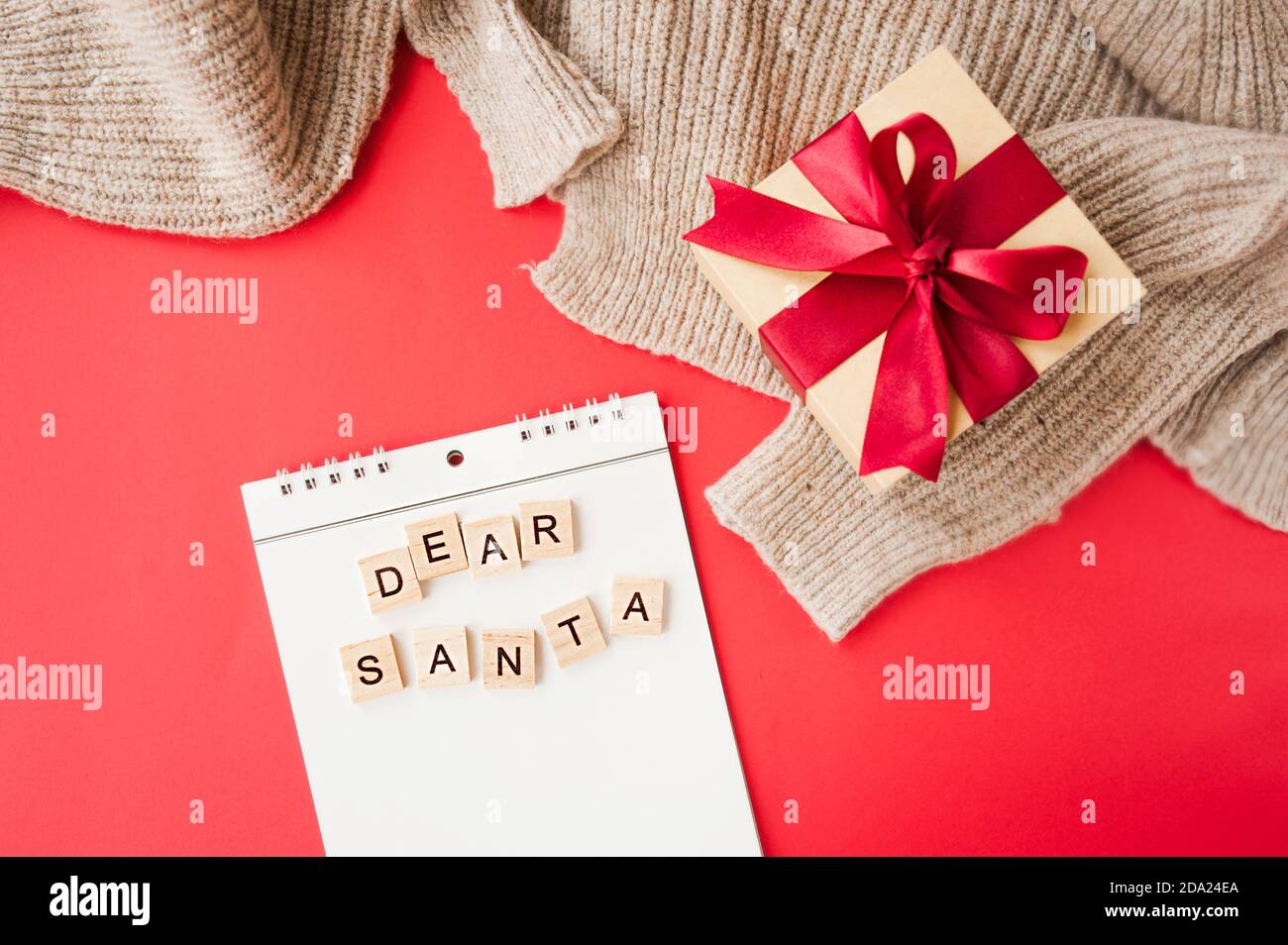 Weißes Spiralheft und quadratische Inschrift lieber Weihnachtsmann auf rotem Hintergrund, Geschenkbox aus Kraftpapier mit roter Satinschleife, Girlanden und gestricktem Fabri Stockfoto