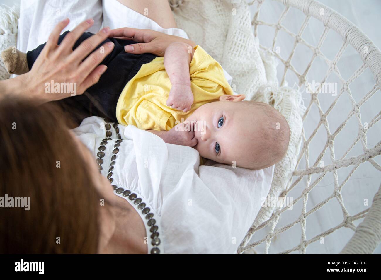Porträt eines Babys mit ausdrucksstarken Augen in einem gelben T-Shirt mit Mama in den Armen in einem Hängemattenstuhl auf weißem Hintergrund Draufsicht. Stockfoto
