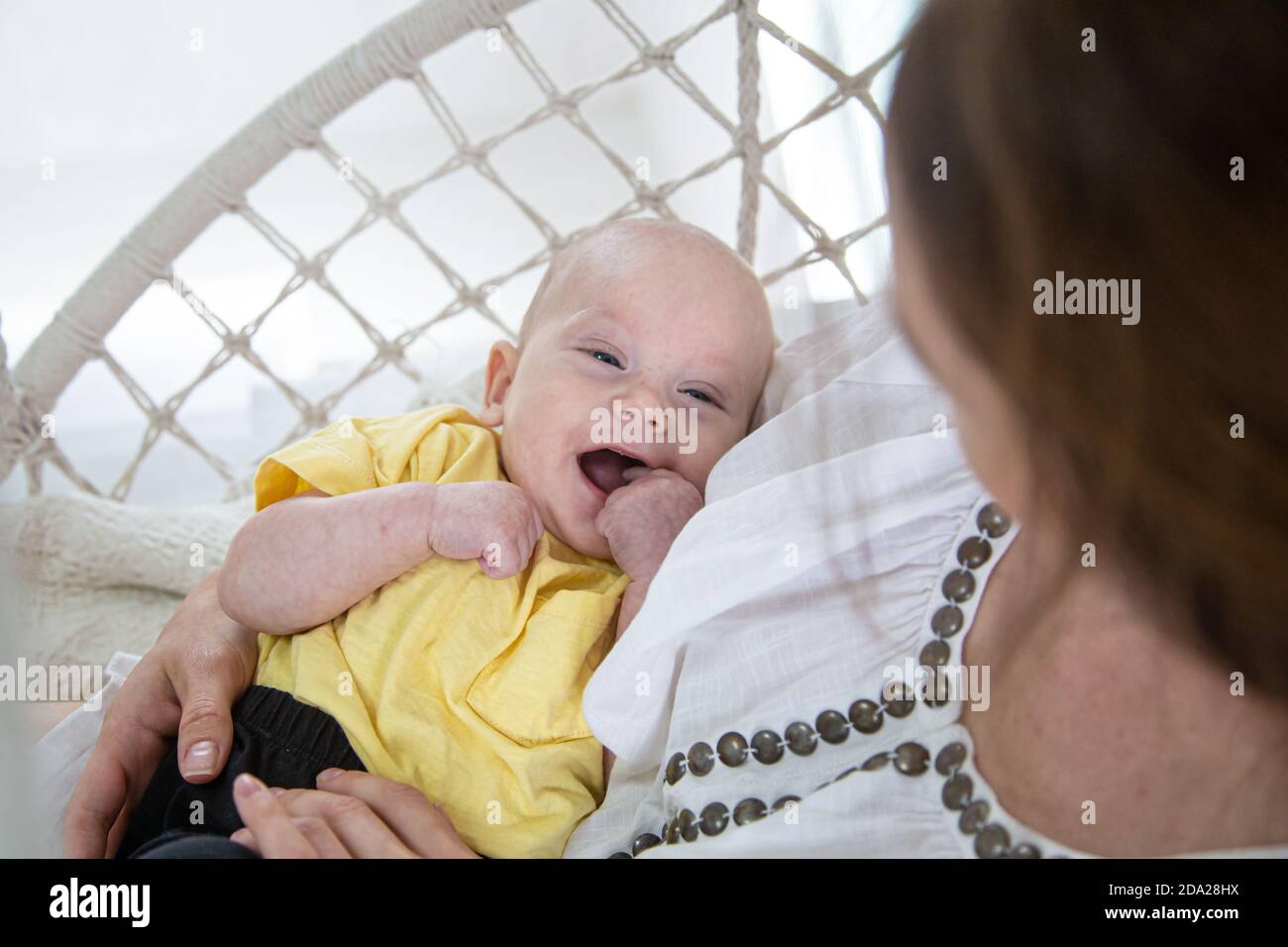 Porträt eines lächelnden Babys in einem gelben T-Shirt mit Mama in ihren Armen in einem Hängemattenstuhl auf einem weißen Hintergrund Kopie Raum. Stockfoto