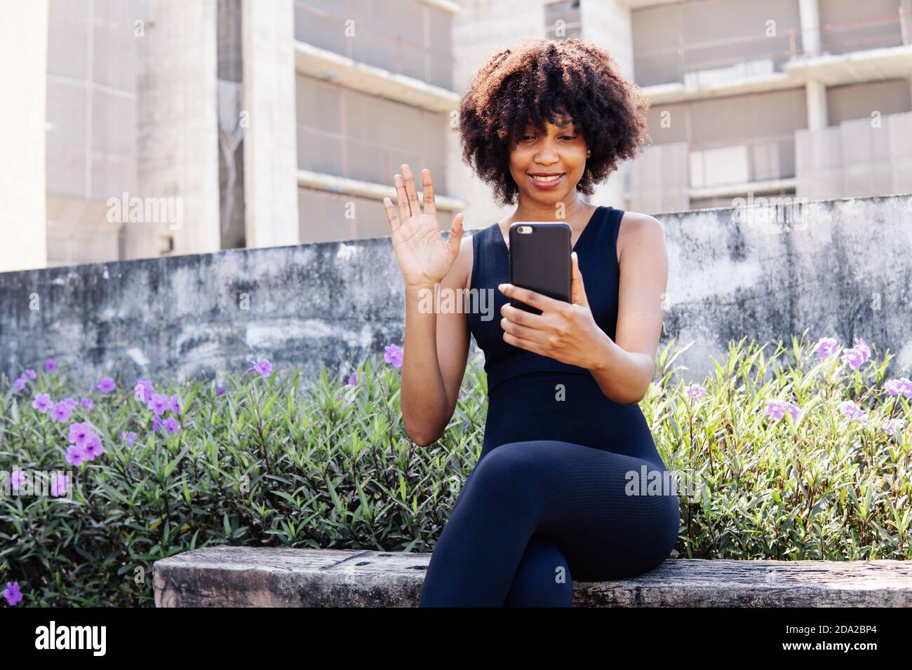 Junge afroamerikanische Frau, die ein Mobiltelefon benutzt und einen Videoanruf macht, während sie auf einer Bank im Park sitzt. Weibliche schwarze Joggerin, die sich ausruhte und die Hände bei der Kamera im Freien winkte Stockfoto