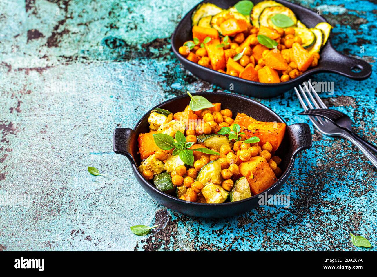 Gebackene Süßkartoffeln, Zucchini und Kichererbsen in gusseisernen Pfannen, blauer Hintergrund. Gebackenes Gemüse. Gesunde Ernährung Konzept. Stockfoto