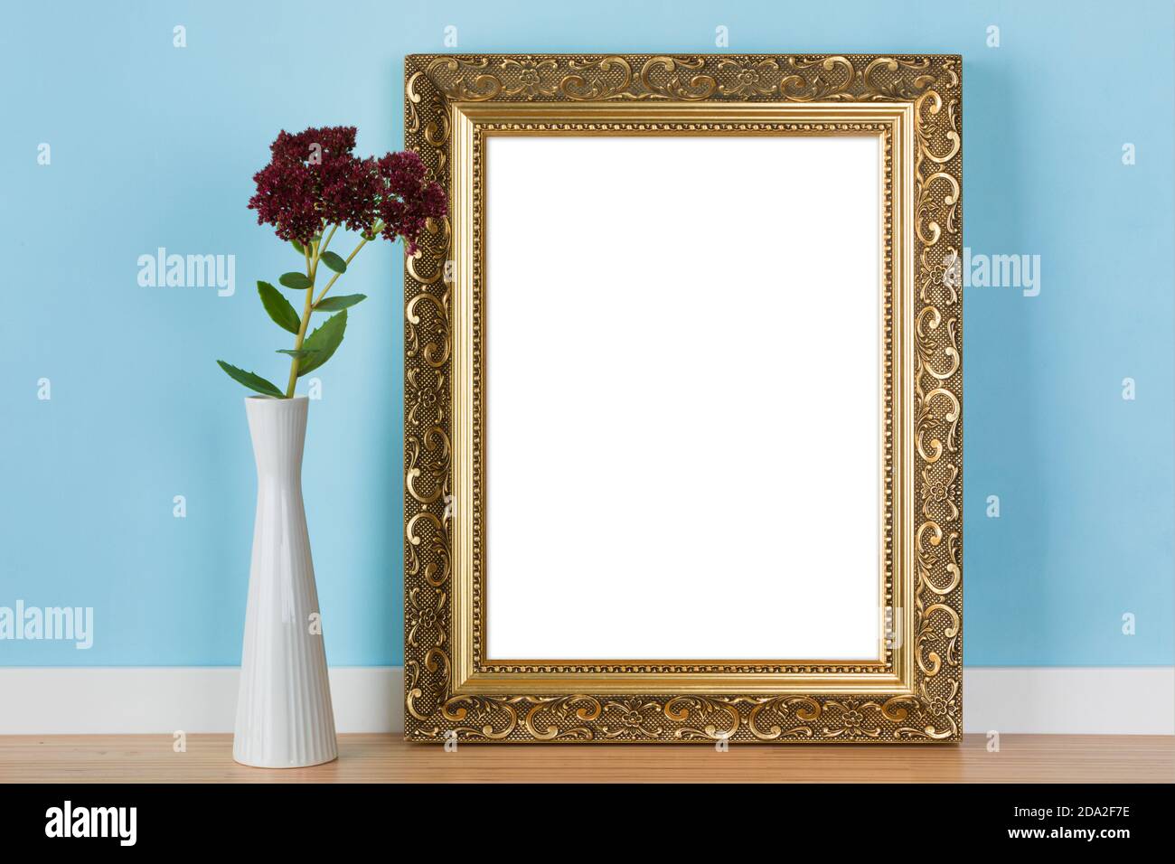 Vertikaler goldener Bilderrahmen an blauer Wand mit Steinbrotblumen in Vase Mockup Schablone gelehnt. Leerer Bildbereich mit Beschneidungspfad maskiert Stockfoto