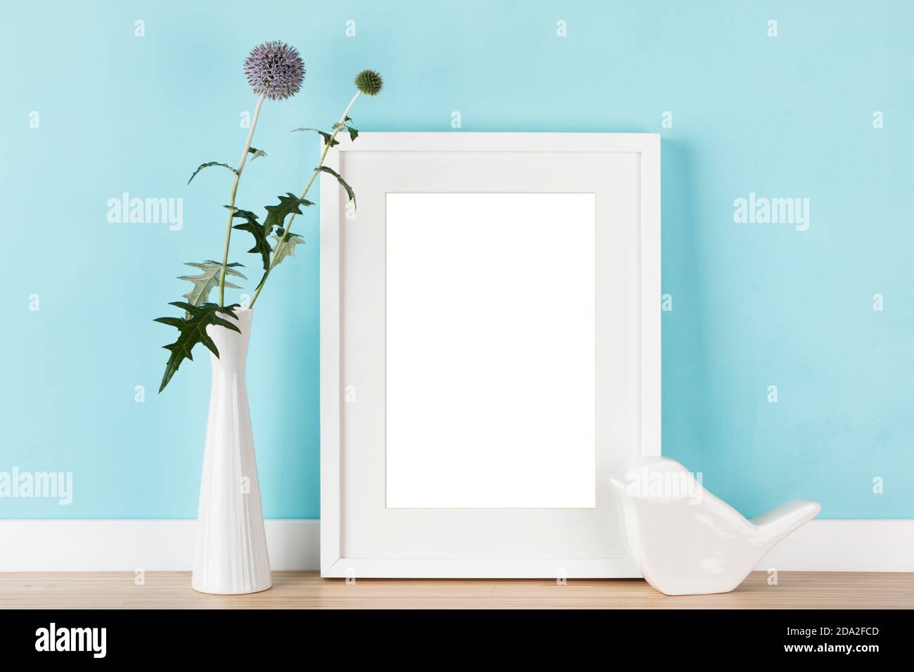 Eleganter vertikaler weißer Bilderrahmen mit mattem Mockup, Schablone mit Kugeldisteln in Vase und stilisiertem Keramikvogel vor pastellblauer Wand. B Stockfoto