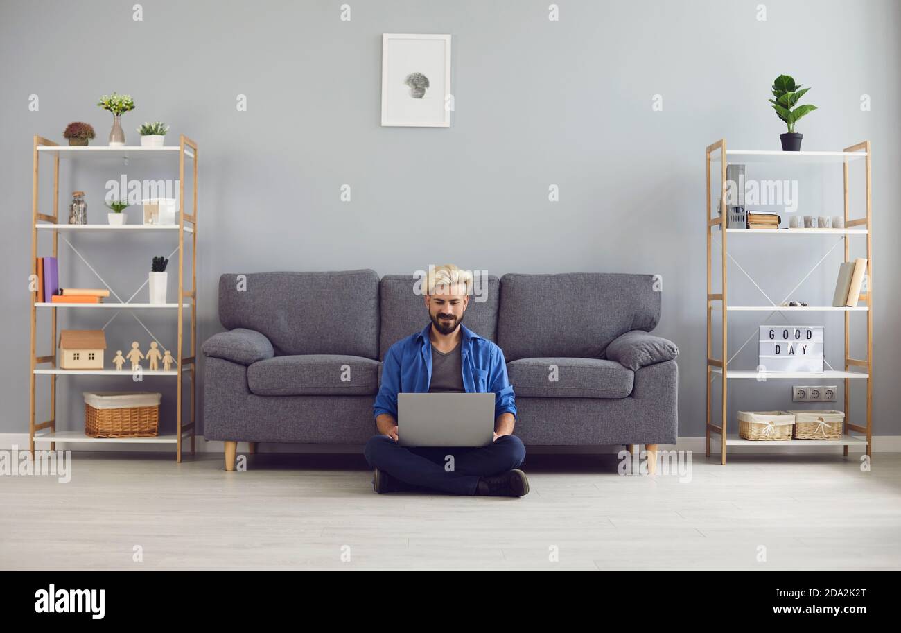 Junger lächelnder Mann, der auf dem Boden sitzt und einen Laptop auf den Knien hat Und von zu Hause aus online arbeiten Stockfoto