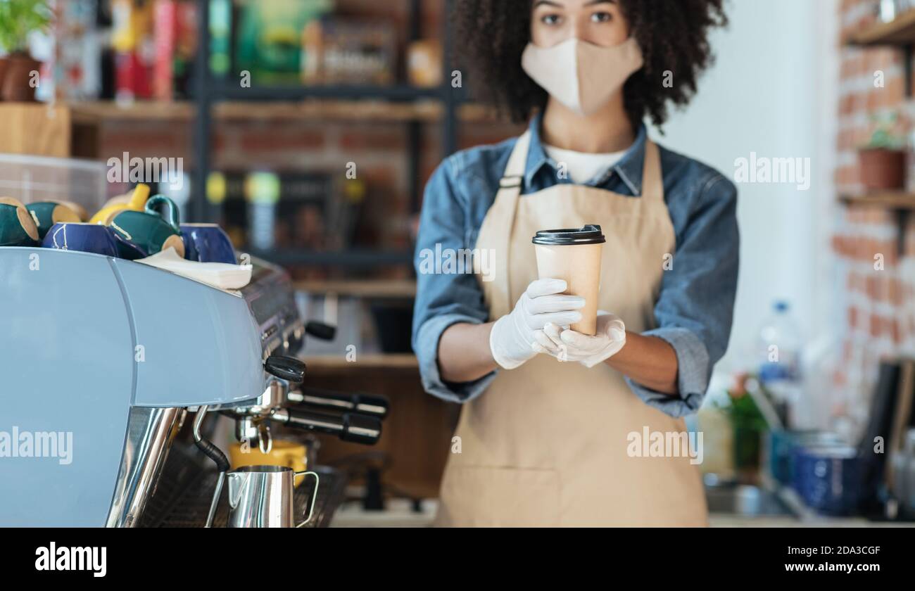 afroamerikanische Hipster Kellnerin in Gesichtsmaske und Handschuhen Hält die Bestellung von Getränken zum Mitnehmen in den Händen, um im Café eine Tasse zu geben Stockfoto