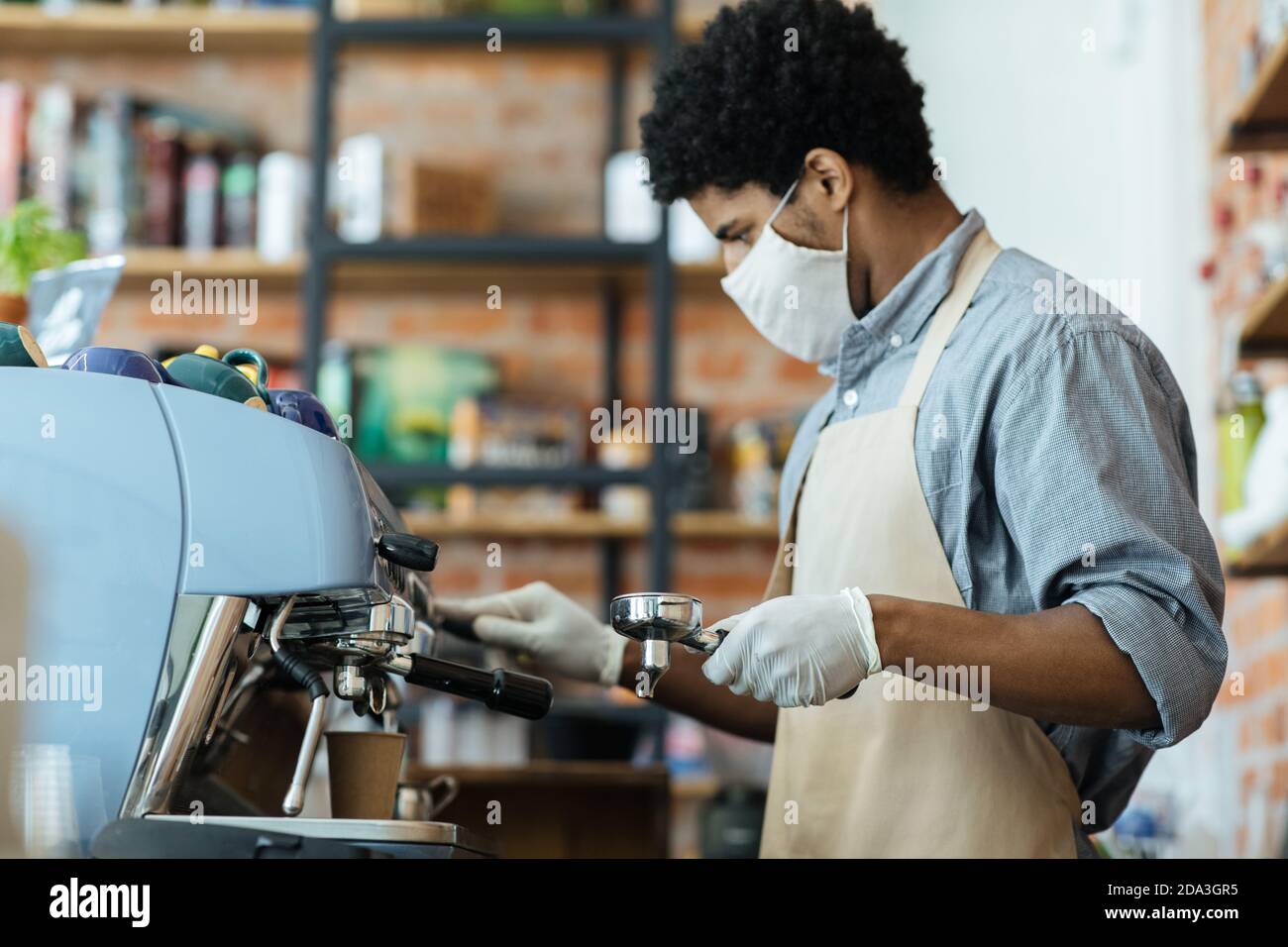 Attraktive Millennial african american Mann in Schürze mit Handschuhen und Schutzmaske macht Espresso Stockfoto