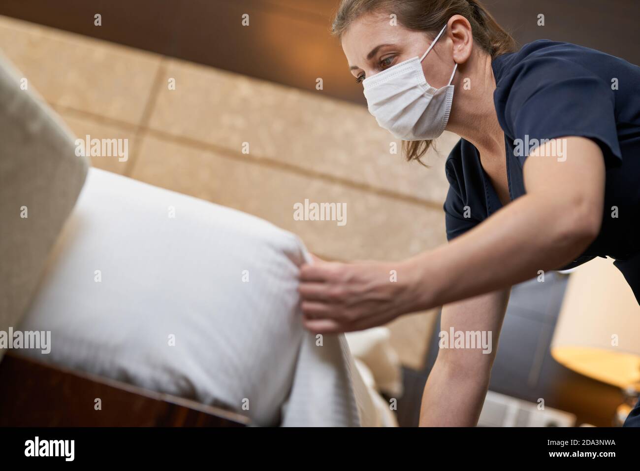 Beschnittenes Foto von der Hotelkammerin, die Uniform und Maske trägt, während sie das Gästebett macht. Servicekonzept des Hotels Stockfoto