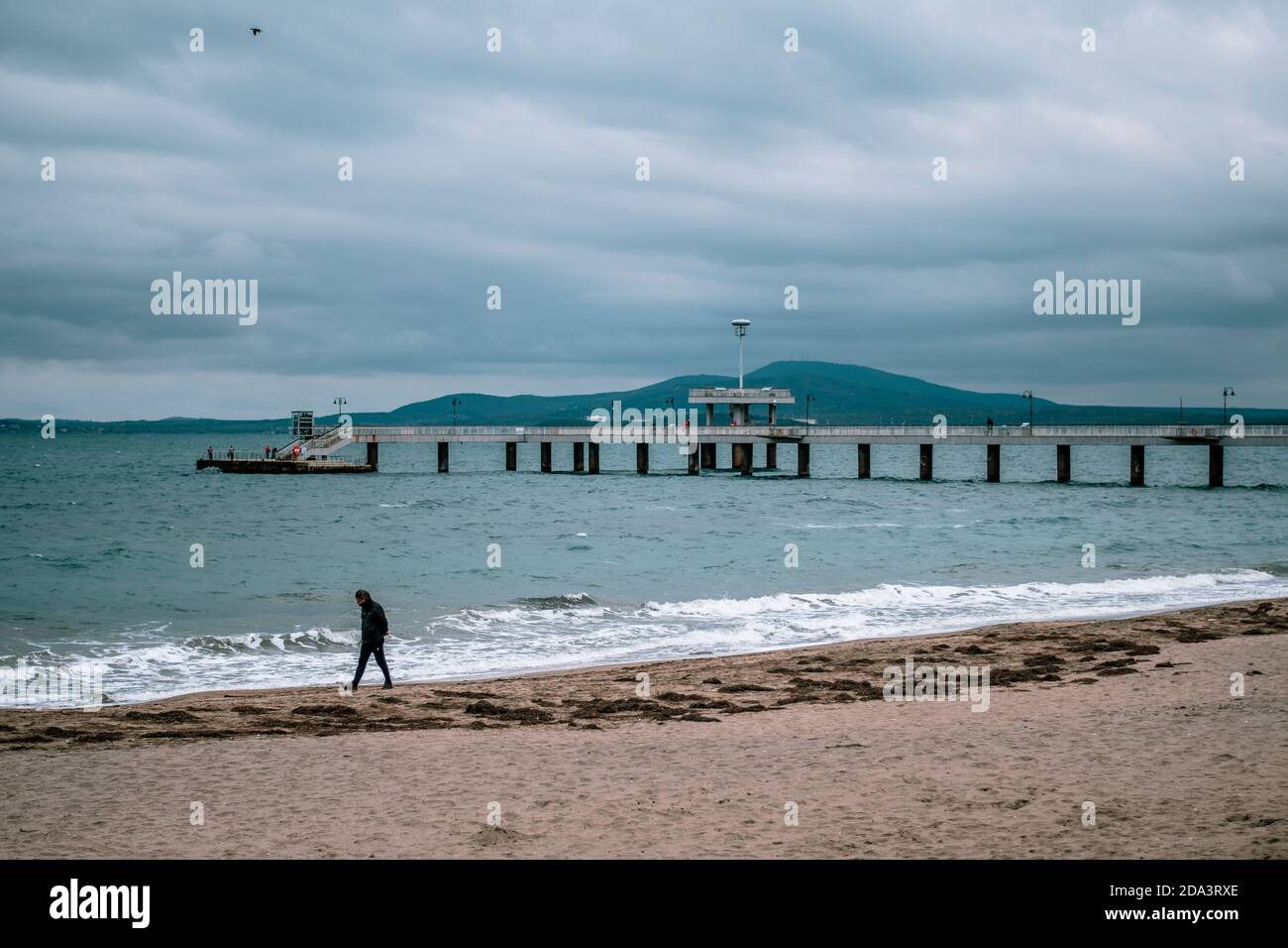 Eine Person, die alleine an einem leeren Strand in Burgas, Bulgarien, läuft. Winter Herbstsaison, kaltes Wetter. Wolkiges Wetter, Seebrücke im Hintergrund. Hohes q Stockfoto