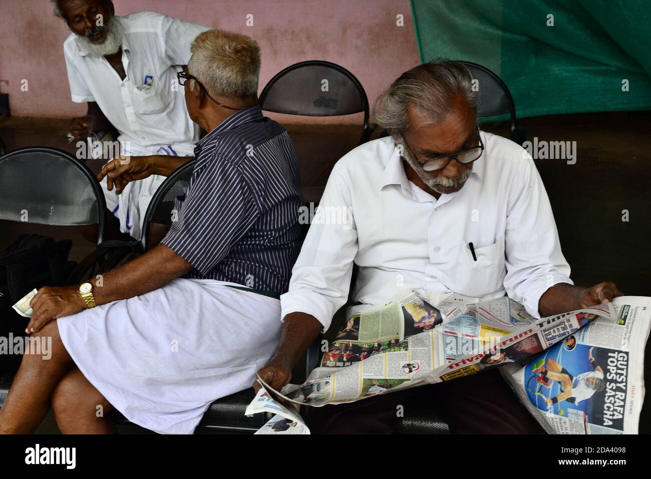 Varkala, Kerala, Indien - Januar, 2017: Alte Haupt in den Gläsern Zeitung auf dem Bahnhof lesen. Zwei alte indische Männer sitzen hinter und reden Stockfoto
