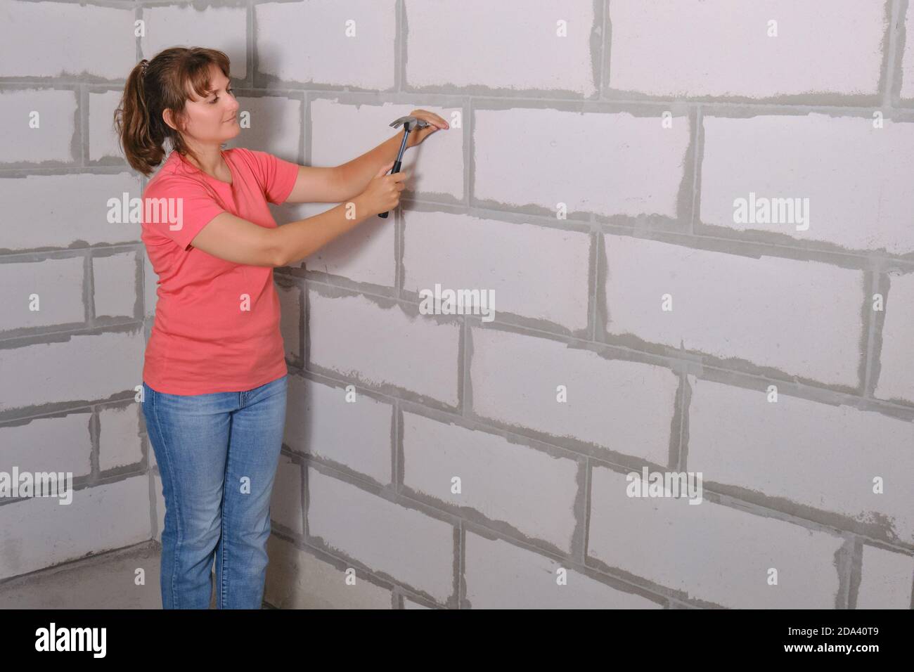 Eine junge Frau mit einem Hammer hämmert einen Nagel in die Wand aus Schaumstoffblöcken. Reparieren Sie mit Ihren eigenen Händen in einer neuen Wohnung, kopieren Raum. Die Schaffung von Stockfoto