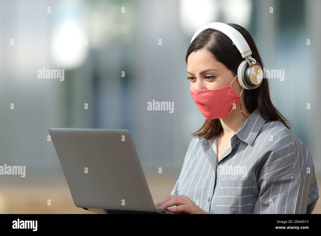Glückliche Frau mit Maske mit Laptop und Kopfhörer sitzen in Die Straße Stockfoto