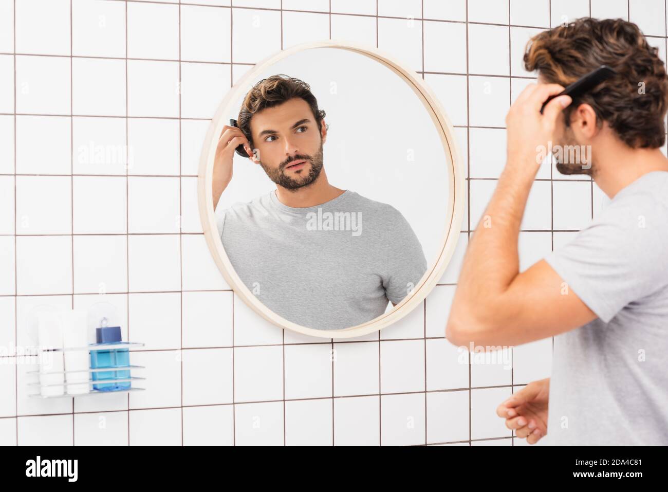 Junger Mann, der die Kamera anschaut, während er die Haare verschwommen kämmt Vordergrund im Badezimmer Stockfoto