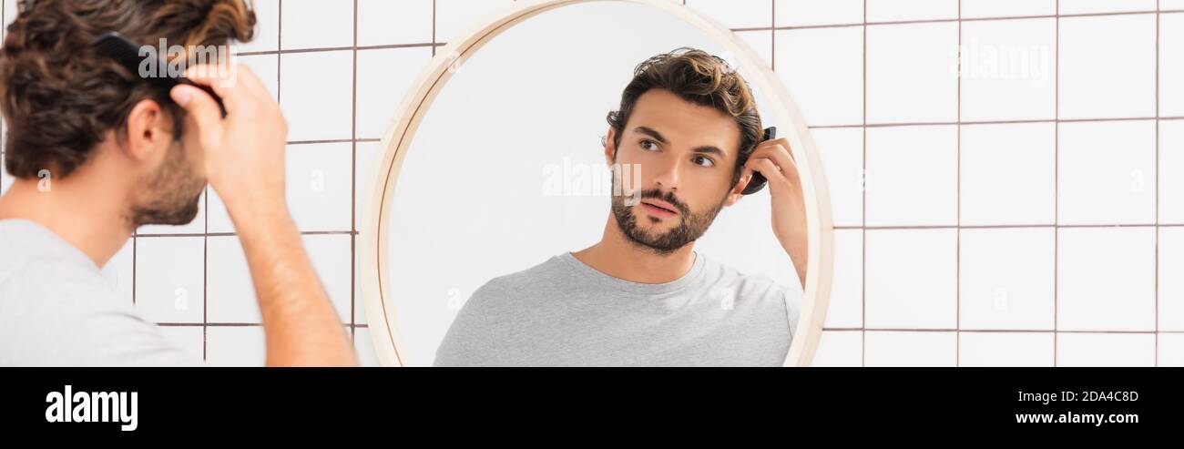 Junger Mann kämmt Haare, während Blick auf Spiegel auf verschwommenem Vordergrund im Badezimmer, Banner Stockfoto