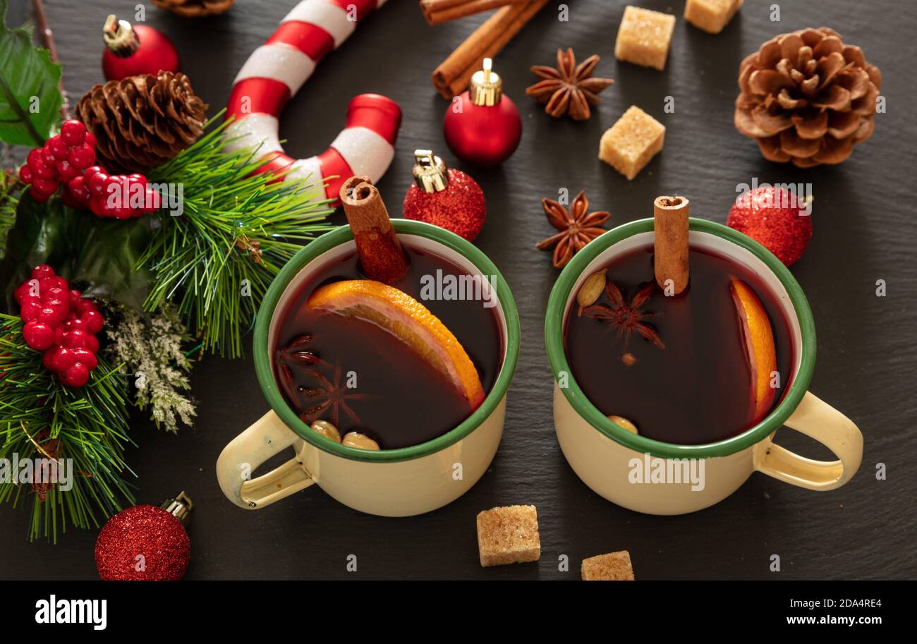 Glühwein, winterwärmendes Alkoholgetränk, zwei Jahrgangsbecher, Zutaten Orange und Gewürze und Weihnachtsdekoration auf schwarzem Stein, Nahaufnahme. Stockfoto