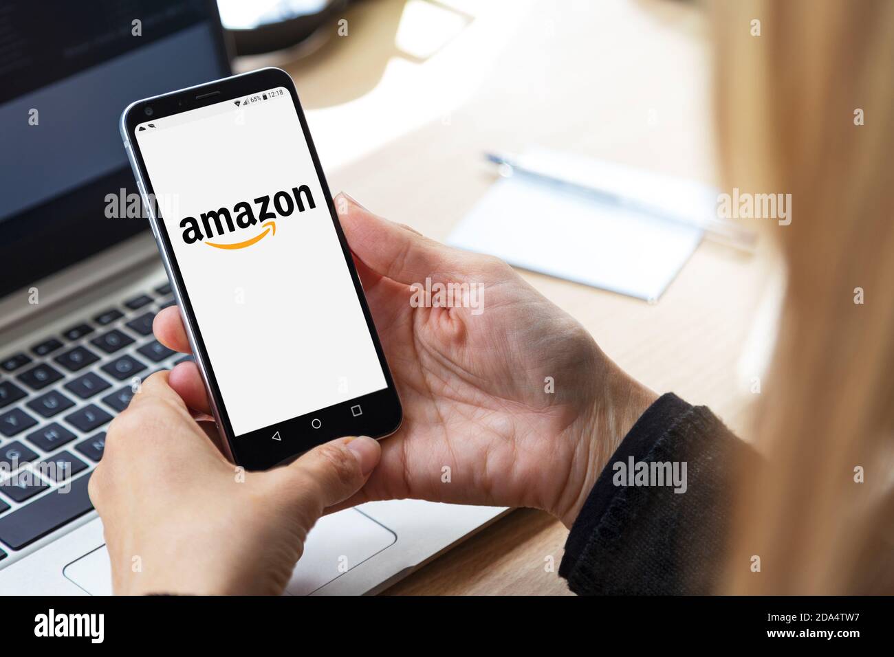 mazon-Logo auf dem Bildschirm des Mobiltelefons. Blonde Frau an ihrem Arbeitsplatz, vor dem Laptop mit Smartphone in Stockfoto