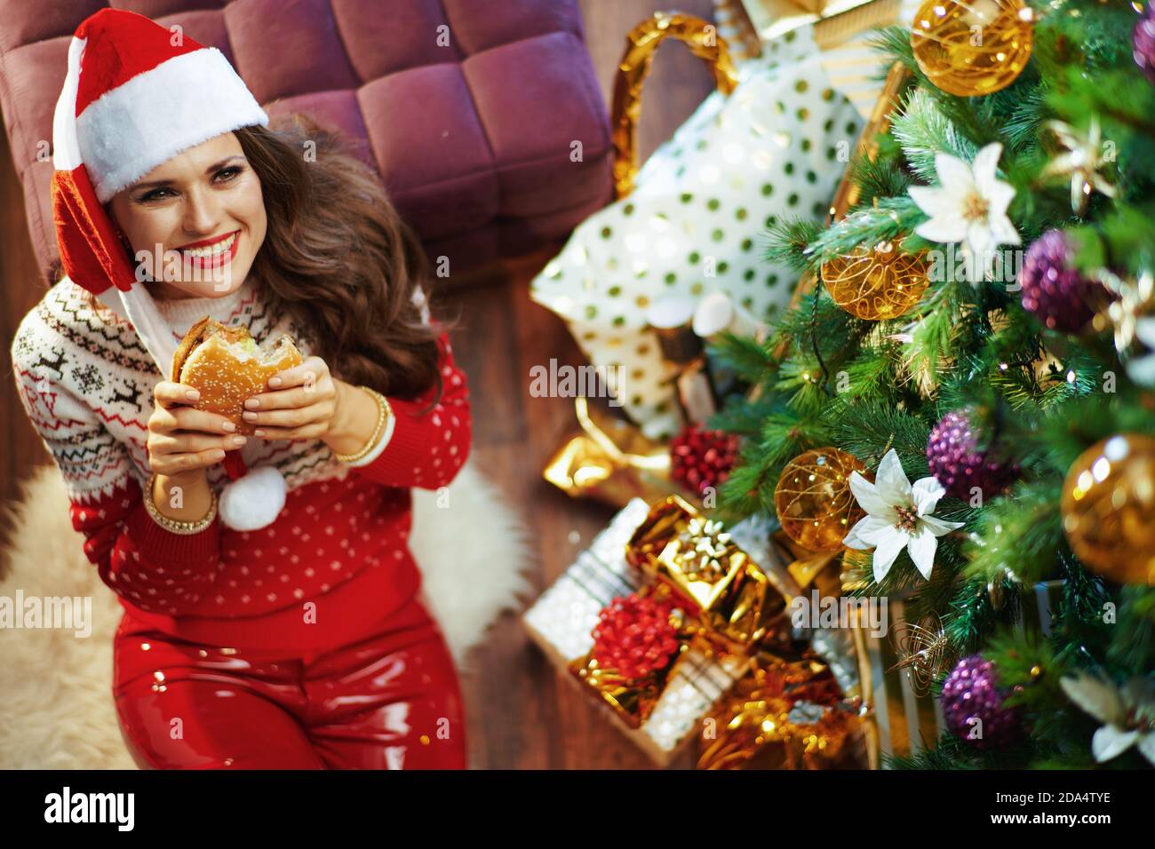 Frohe Weihnachten. Obere Ansicht der glücklichen eleganten Frau in roten Pullover und Weihnachtsmann Hut in der Nähe von Weihnachtsbaum und Geschenkboxen Essen Hamburger. Stockfoto