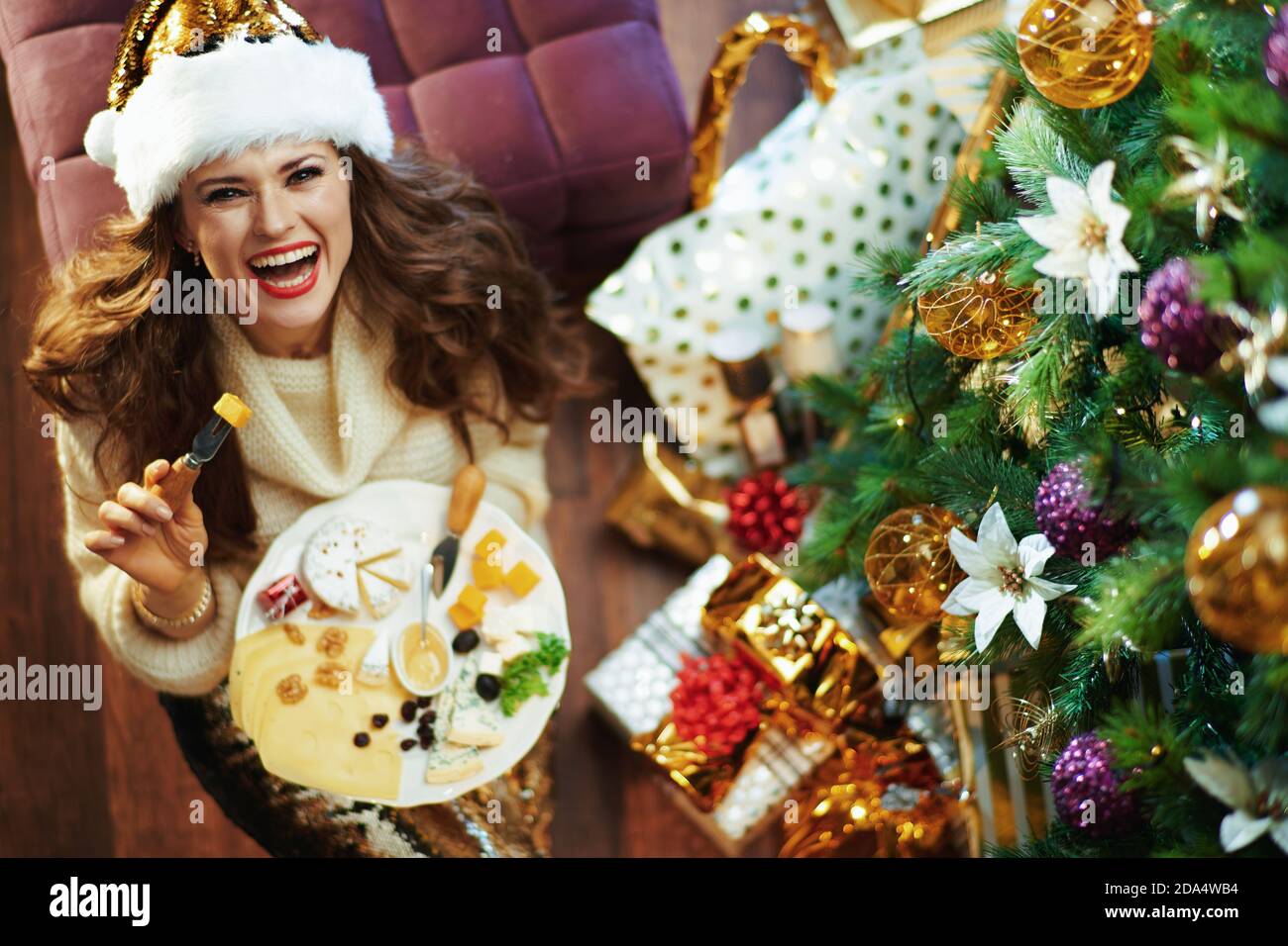 Frohe Weihnachten. Obere Ansicht der lächelnden 40 Jahre alte Hausfrau in Gold Pailletten Rock und Pullover mit großen Käseplatte in der Nähe von Weihnachtsbaum und Geschenk-Box Stockfoto