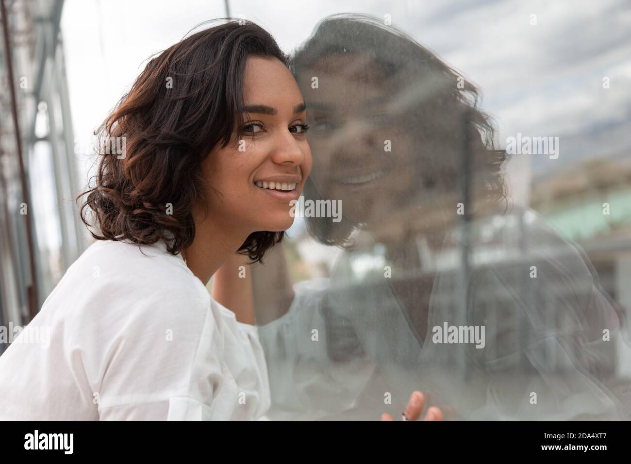 Fröhliche Person mit seinem Gesicht steht neben ihrem Spiegelbild in einem Spiegel, hat eine weiße Bluse, im Freien mit modernem Stil Stockfoto