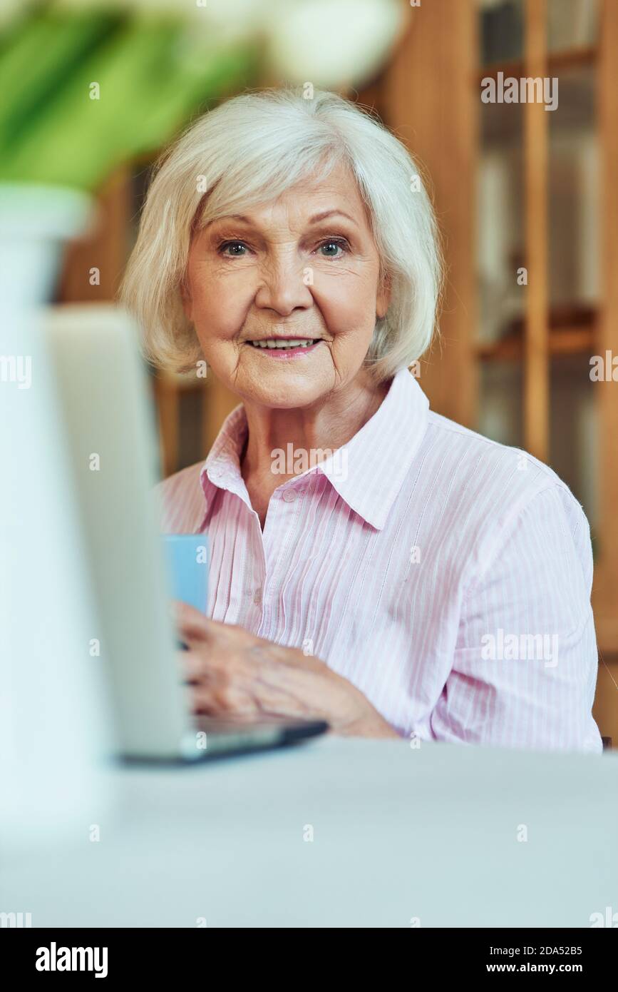 Glückliche ältere Frau, die morgens Kaffee genießt, während sie zu Hause sitzt und Laptop benutzt. Inländisches Lifestyle-Konzept Stockfoto