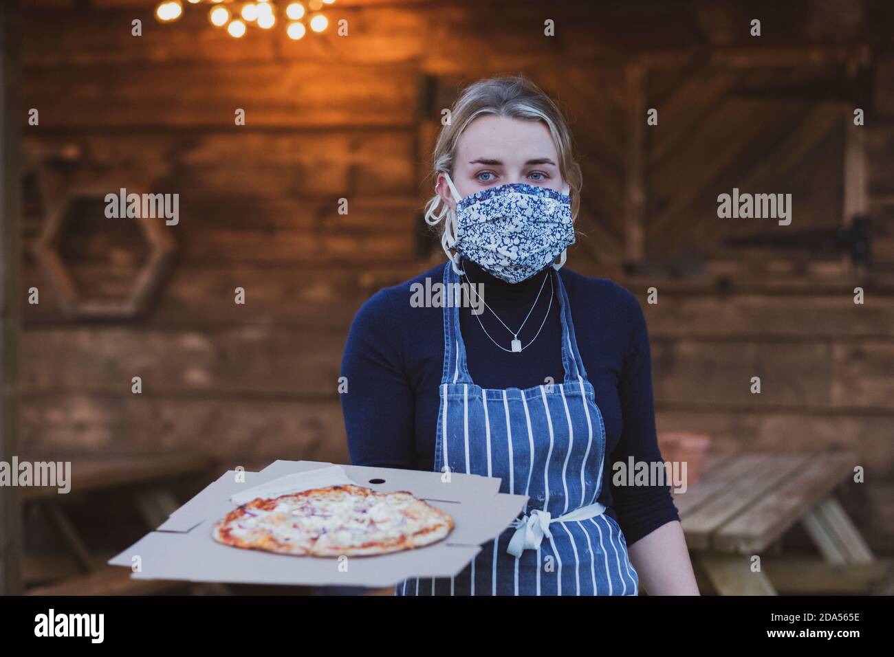 Kellnerin in Schürze und Gesichtsmaske hält Teller Pizza. Stockfoto