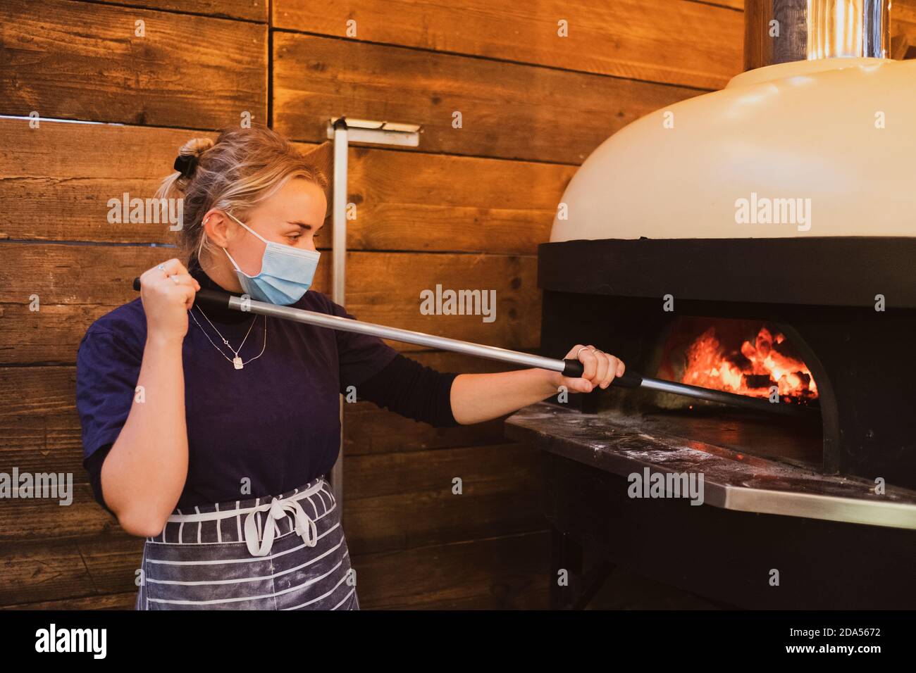 Blonde Kellnerin trägt blau gestreifte Schürze und Gesichtsmaske stehen an einem Pizzaofen in einem Restaurant. Stockfoto