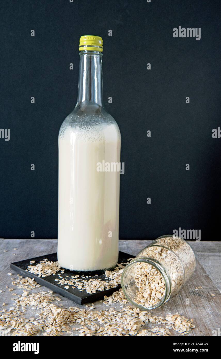 Vegane Hafermilch in hoher Flasche mit Hafer auslaufen Von Glas auf schwarzer Kachel vor schwarzem Hintergrund Stockfoto