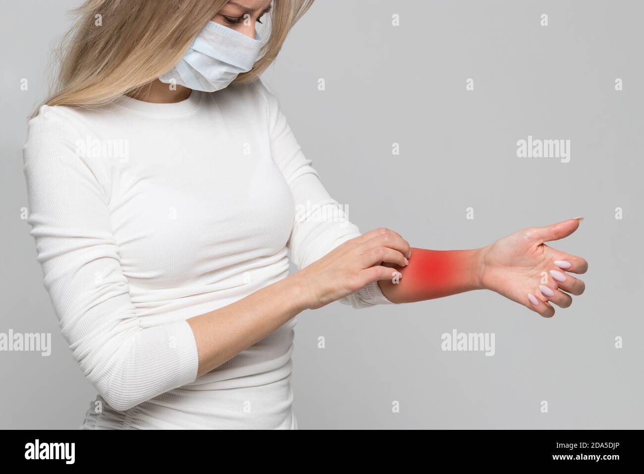 Studio Porträt der Frau in medizinischen Gesichtsmaske kratzenden Arm in rot, isoliert auf grauem Hintergrund. Symptome einer Tierallergie, juckende Haut Stockfoto