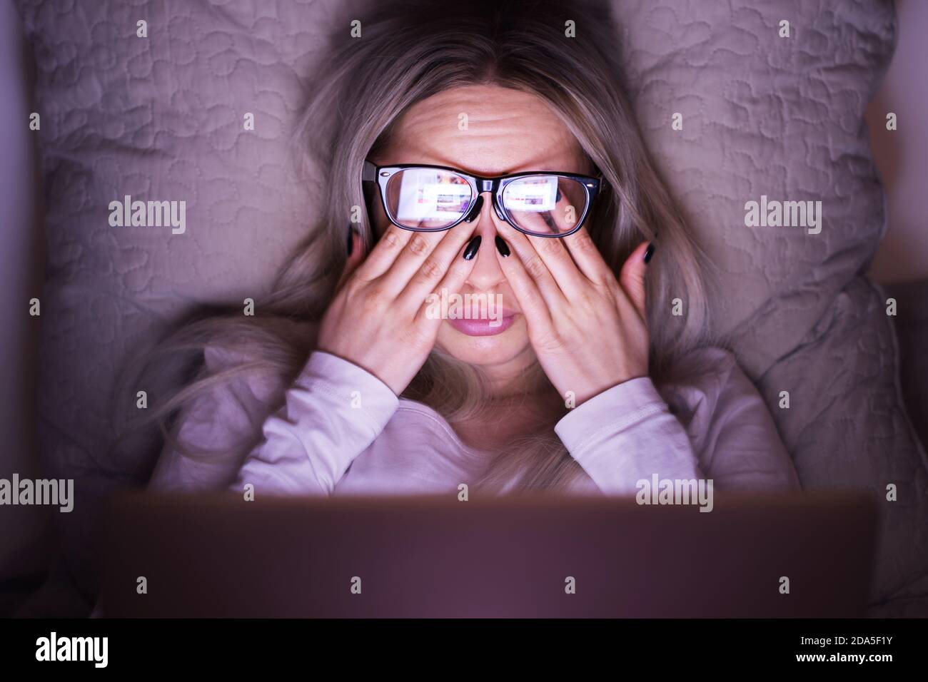 Schläfrige junge kaukasische Frau in einer Brille, die ihre Augen reibt, fühlt sich müde nach der Arbeit an einem Laptop, auf dem Kissen liegend, Vorderansicht/ Überarbeit, müde, Gesundheit Stockfoto