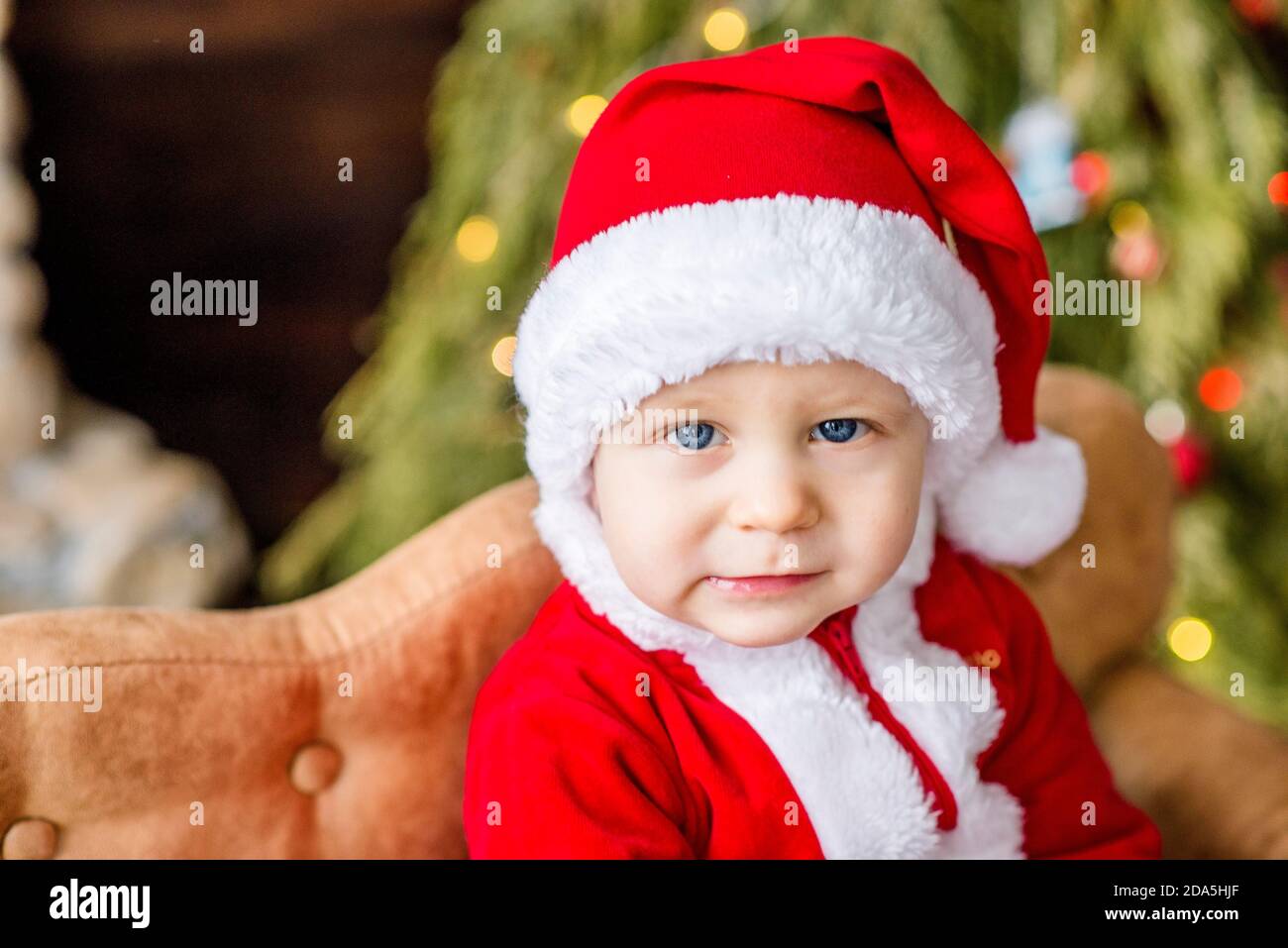 Nahaufnahme Porträt eines kleinen Jungen als roter Weihnachtsmann gekleidet, spielen am Steinkamin mit hellen Girlanden, setzen eine Geschenkbox unter Weihnachtsbaum Stockfoto