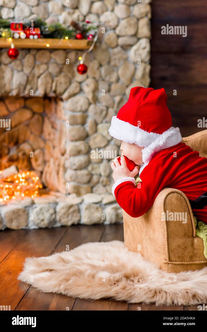 Ein kleines Kind im roten Weihnachtsmann Kostüm, spielt in seiner Residenz, leckt einen Neujahrsball wie Süßigkeiten. Festliche Postkarte. Der blonde Junge lacht. Weihnachten Stockfoto