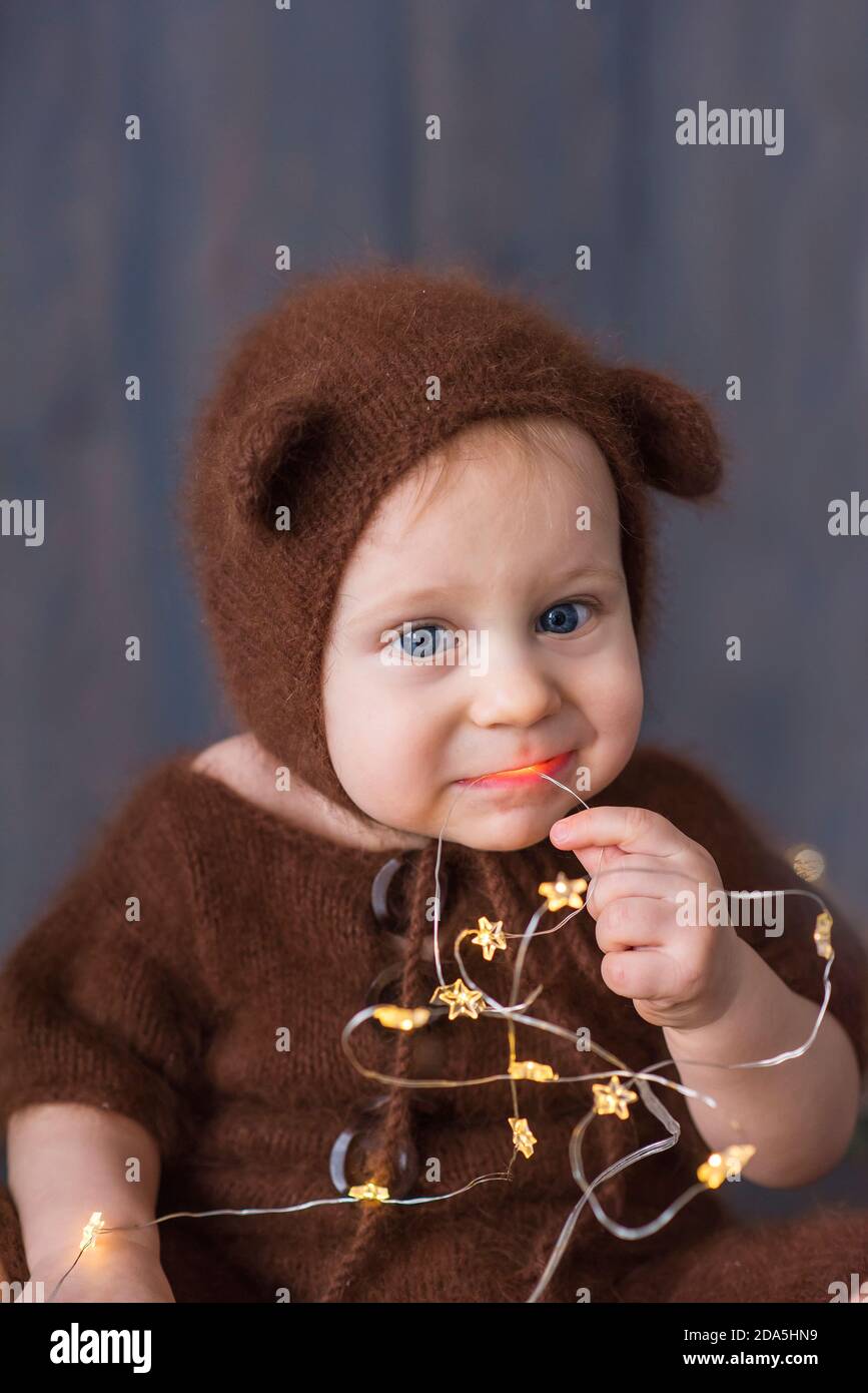 Fröhlicher kleiner Junge in einem braunen, flauschigen, gestrickten Bärenkostüm, sitzt auf dem furon auf dem Holzboden, spielt mit einer hellen weihnachtlichen Lichtergirlande. Stockfoto