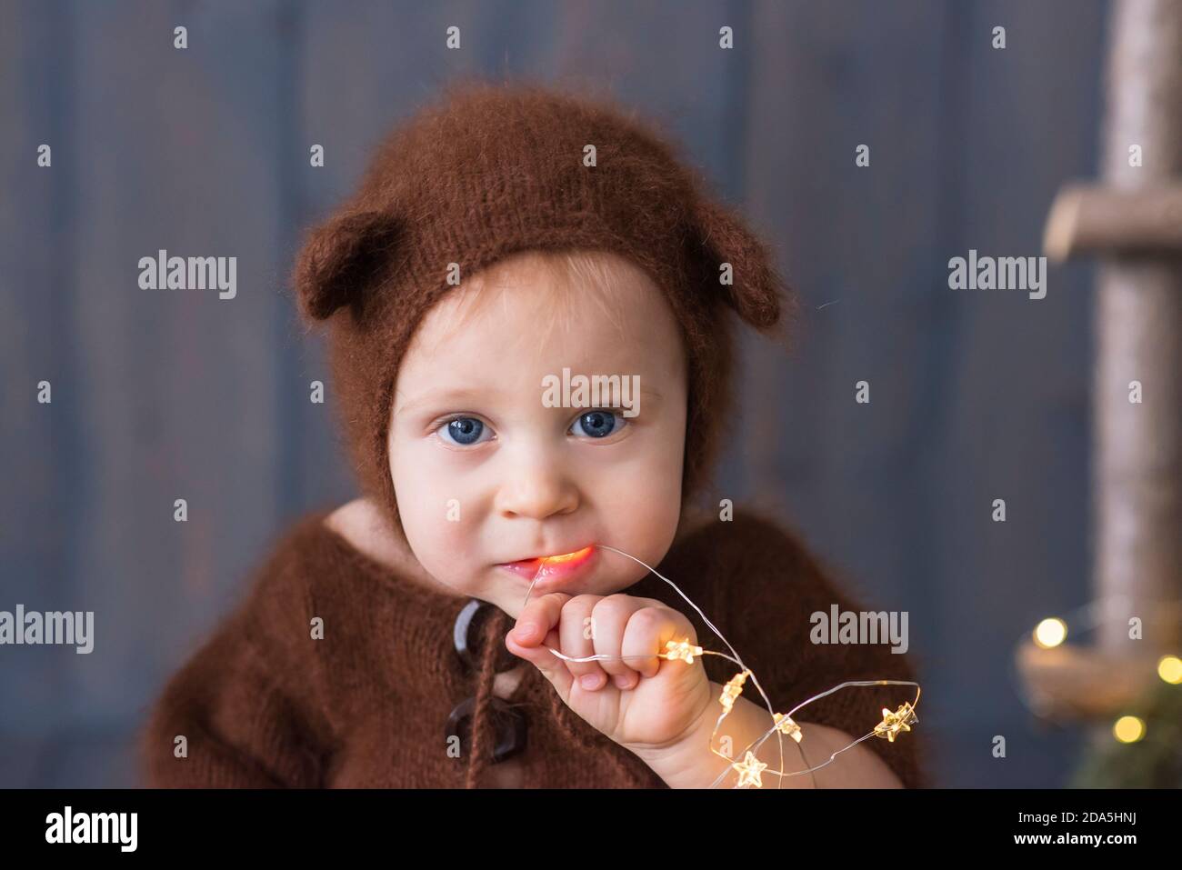 Fröhlicher kleiner Junge in einem braunen, flauschigen, gestrickten Bärenkostüm, sitzt auf dem furon auf dem Holzboden, spielt mit einer hellen weihnachtlichen Lichtergirlande. Stockfoto