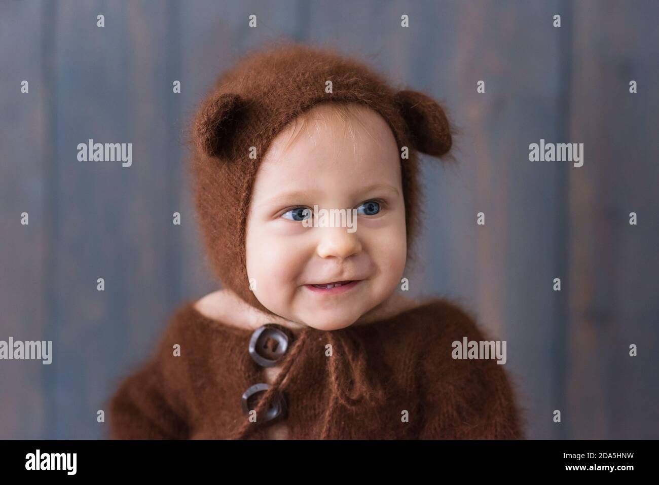 Fröhlicher kleiner Junge in einem braunen, flauschigen, gestrickten Bärenkostüm, sitzt auf dem furon auf dem Holzboden, spielt mit einer hellen weihnachtlichen Lichtergirlande. Stockfoto