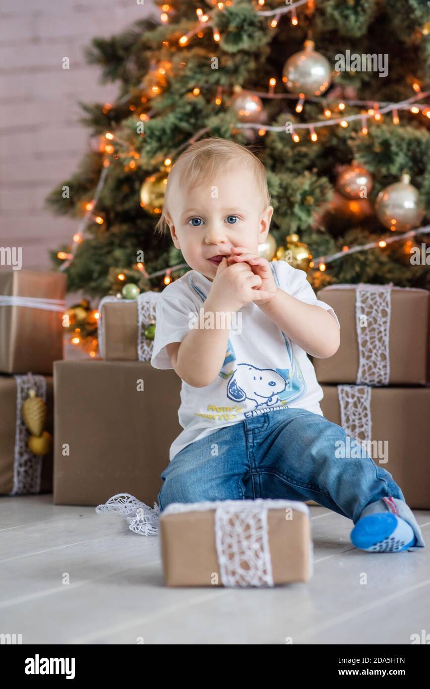 Kleiner blonder Junge, der einen Lebkuchenmann am Weihnachtsbaum zu Hause isst. Neujahrskarte mit Girlanden Bokeh. Das Baby lacht, öffnet eine Geschenkbox Stockfoto